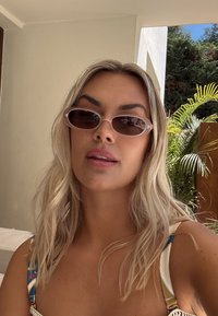 Szőke nő ovális napszemüveget visel és mintás ujjatlan felsőt, beltéren pózol egy ajtó közelében, ahol kívül növények láthatók.