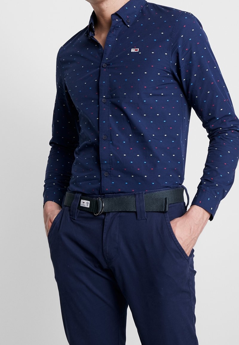 Homme portant une chemise à boutons bleu marine avec de petits points multicolores et un pantalon bleu marine assorti avec une ceinture noire, les mains dans les poches.