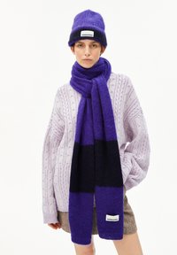 Femme portant une écharpe et un bonnet rayés violet et noir, avec un pull en tricot à torsades violet clair et une jupe marron sur fond blanc.