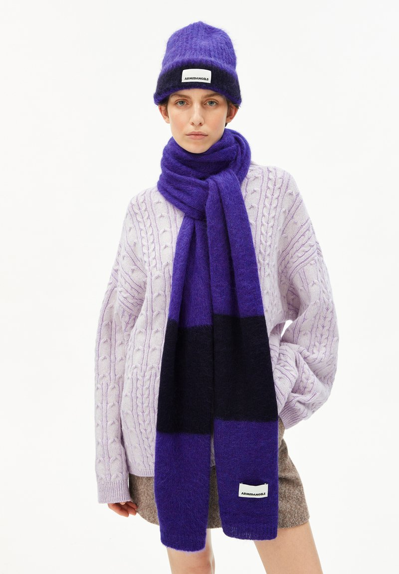 Femme portant une écharpe et un bonnet rayés violet et noir, avec un pull en tricot à torsades violet clair et une jupe marron sur fond blanc.