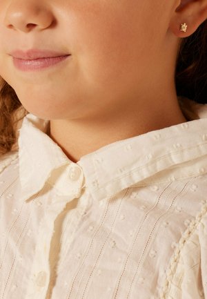 L'enfant porte un chemisier blanc texturé avec un col à boutons. Il présente des points en relief et des rayures subtiles ; une petite boucle d'oreille en or est visible.