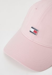 Gorra de béisbol rosa de algodón con una superficie texturizada, que presenta un logo rectangular de "Tommy Jeans" en la parte frontal.