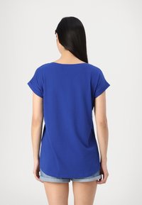 Pitkähiuksinen nainen, jolla on sininen lyhythihainen paita ja käärityt farkkushortsit, selin kirkkaan vaalean taustan edessä.