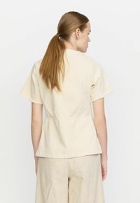 Beige bomullstopp med korta ärmar och fransade kanter samt ett peplum-snitt. Bakom finns en söm som går vertikalt ner längs mitten.