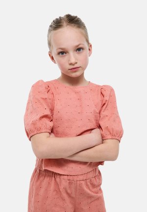 Jeune fille aux cheveux clairs tirés en arrière, portant un haut rose à motif floral et un bas assorti, les bras croisés, avec une expression neutre.