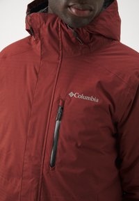 Rød Columbia-jakke med høj krave, lynlåslomme på brystet og tekstureret stof med vandrette linjer.