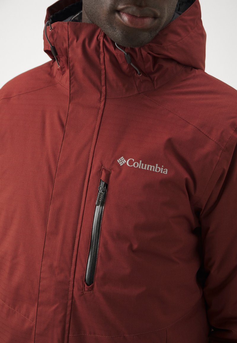 Chaqueta roja de Columbia con cuello alto, bolsillo en el pecho con cremallera y tejido texturizado con líneas horizontales.