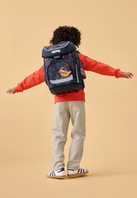 Zaino ergonomico nero e rosso con motivo, con spallacci regolabili, forma curva, dettagli riflettenti e adesivi circolari. Indossato da un bambino.