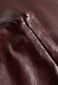 Burgundska teksturirana umetna snov z sijočo površino, ki ima zadrgo ob enem robu in zguban izgled.