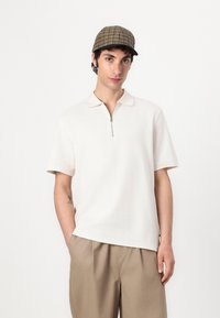 Cream short-sleeve polo shirt with a zippered collar, paired with light brown trousers. The model also wears a check-patterned cap.