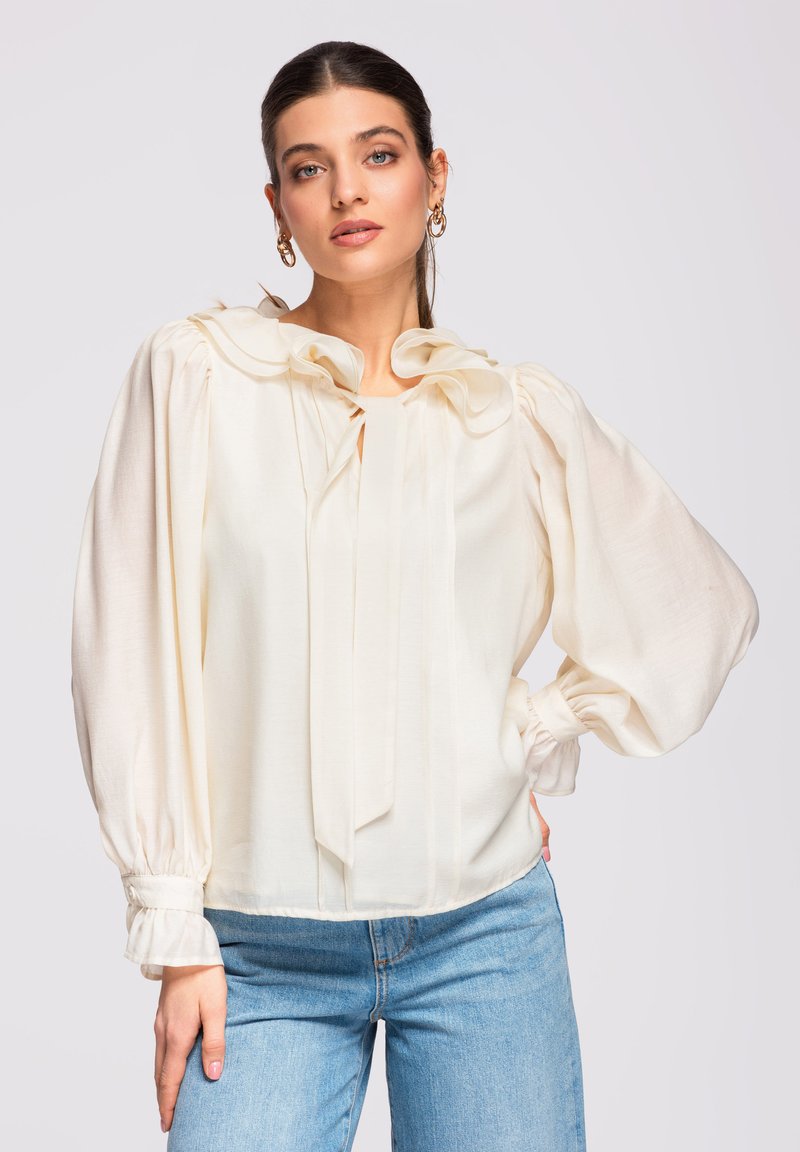 Femme aux cheveux foncés portant des boucles d'oreilles créoles dorées, un chemisier crème à manches bouffantes et col à volants, ainsi qu'un jean bleu clair, posant avec la main sur la hanche.