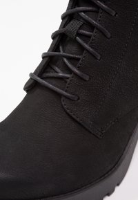 Gros plan sur une botte en cuir noir avec des lacets ronds et une surface texturée, montrant des coutures détaillées et une semelle épaisse sur fond blanc.