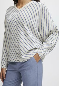 Maglione a V in crema con strisce diagonali blu. Vestibilità ampia con maniche lunghe. Abbinato a pantaloni su misura blu chiaro.
