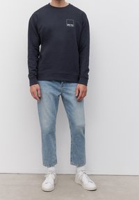 Homme portant un sweat-shirt bleu marine avec le logo "MO'PD", un jean droit bleu clair, des chaussettes blanches et des baskets blanches, debout contre un mur blanc.