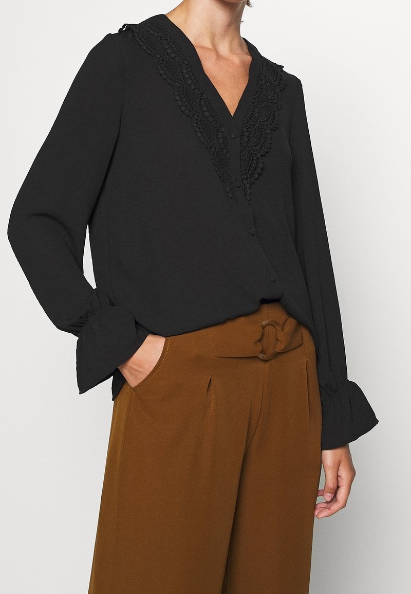 Blouse noire avec des détails en dentelle le long de l'encolure, à boutons sur le devant, et manches longues avec des poignets froncés, associée à un pantalon brun taille haute.
