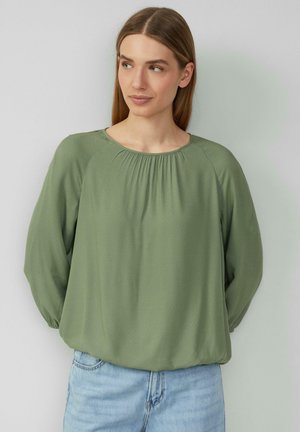 Hellgrüne Bluse mit rundem Ausschnitt, Raffung und langen Ärmeln. Weicher Stoff und lockere Passform, kombiniert mit hellblauen Jeans.
