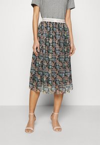 Kobieta nosząca plisowaną, kwiatową spódnicę midi z białym paskiem w talii, na którym napisano "SNY SIĘ SPEŁNIAJĄ, SPEŁNIJ SWOJE", połączoną z beżowymi sandałami na kostkę.