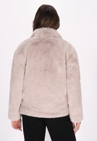 Jachetă din blană sintetică de culoare bej deschis, cu guler, mâneci lungi și croială lejeră. Textură moale, design simplu, fără elemente de fixare vizibile.