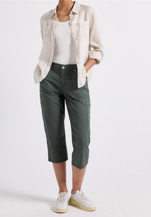 Persoon met opgerolde beige overhemd, wit hemdje, donkergroene cropped broek en witte sneakers, staand met handen in de zakken.