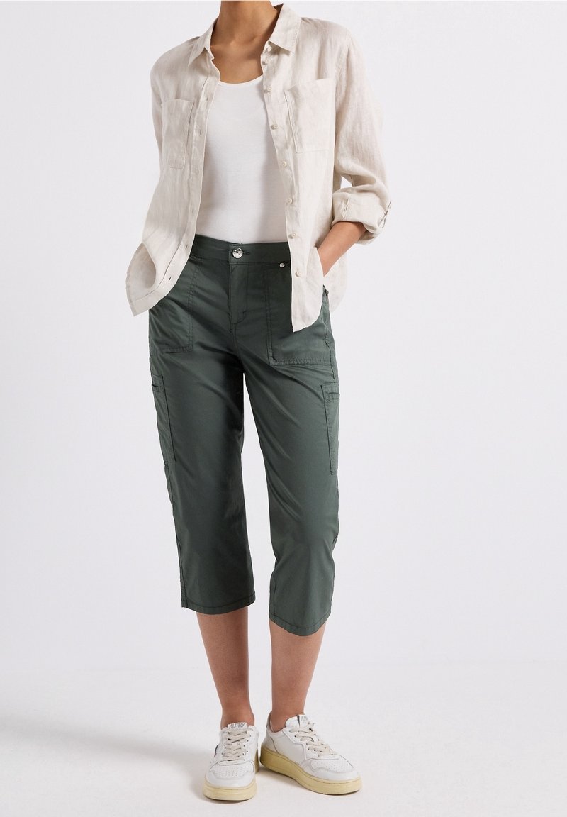 Personne portant une chemise beige retroussée, un débardeur blanc, un pantalon court vert foncé et des baskets blanches, debout les mains dans les poches.