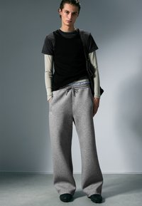 Gray sweatpants, black tank top over a light long-sleeve shirt, combined with dark shoes. Model stands against a light background.