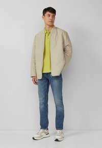 Beżowa pikowana kurtka, zielony polo, niebieskie dżinsy i szare sneakersy z białymi akcentami. Gładka tekstura, luźny krój.