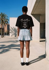 Osoba stoji vani okrenuta leđima, nosi crnu majicu s grafičkim uzorkom, svijetloplavu traper suknju s volanima na rubu, bijele čarape i crne cipele.