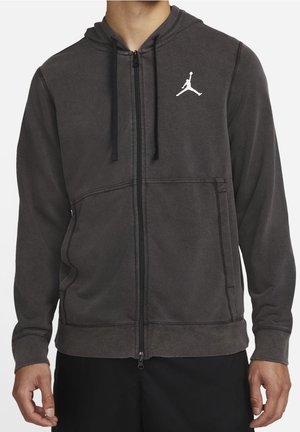 Uomo che indossa una felpa con cappuccio grigio scuro con cerniera, tasche anteriori e logo Jumpman bianco sul petto sinistro.