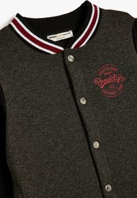 Grå sweatshirt med sorte ærmer, ribbet krave med maroon og hvide striber, metal trykknapper og rød trykt "Brooklyn" logo.
