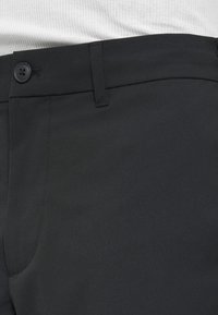 Nærbillede af taljebåndet på sorte bukser med knaplukning og bæltestrop over en hvid ribstrikket top.