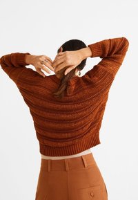 Sweter z dzianiny w kolorze rdzy z teksturowanym wzorem, krótkim krojem i prążkowanymi mankietami, noszony z wysokim stanem brązowych spodni.