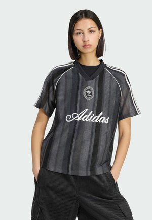 Młoda kobieta o prostych ciemnych włosach, ubrana w czarną sportową koszulkę siateczkową Adidas z białymi paskami i logo, stojąca z rękami w kieszeniach.