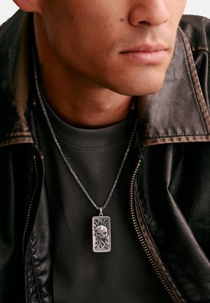 Man wearing a black leather jacket and dark shirt with a silver rectangular pendant necklace featuring a skull and flame design.