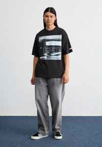 Sort t-shirt med et stort grafisk print i grå og hvid, parret med løstsiddende grå jeans og sorte sko med hvid sål.