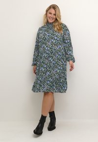 Vestido floral con patrones en negro, azul y verde, mangas largas, puños fruncidos, forma en A y dobladillo a la altura de la rodilla, combinado con botas negras hasta el tobillo.