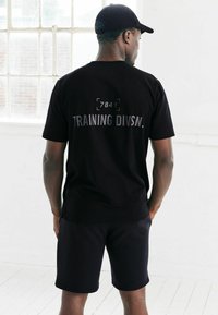 Musta puuvillane T-särk, mille seljal on hallilt trükitud tekst "TRAINING DIVSN." ja number "7841". Kandmine sobivate mustade lühikeste pükstega; mõlemad on lõdva lõikega.