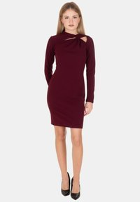 Abito bordeaux a maniche lunghe con scollo annodato e dettaglio a taglio. Linea aderente, tessuto morbido, abbinato a décolleté nere.