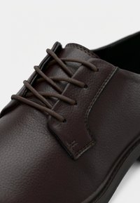 Καφέ δερμάτινο παπούτσι με λείο υφή, στρογγυλή μύτη και καφέ κορδόνια. Σημαντικές λεπτομέρειες περιλαμβάνουν ένα ραμμένο διακοσμητικό στο επάνω μέρος.