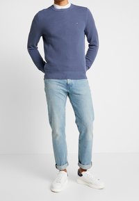 Marinblå stickad tröja med rund halsringning, ribbade manschetter och nederkant, kombinerad med ljusblå jeans och vita sneakers.