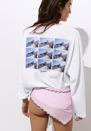 Donna che indossa una camicia bianca a maniche lunghe con stampa a rete di paesaggio di montagna e spiaggia sulla schiena e gonna rosa asimmetrica con bordi smerlati.