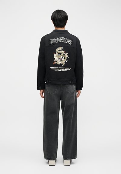 Άτομο που φοράει μαύρο μπουφάν με το κείμενο "MADNESS" και γραφικό φίδι στην πλάτη, σε συνδυασμό με φαρδιά σκούρα γκρι τζιν και λευκά παπούτσια.