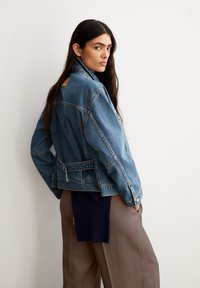 Vrouw met lang, donker haar die een blauwe denimjack en bruin brede broek draagt, staande met handen in de zakken, kijkend over haar schouder.