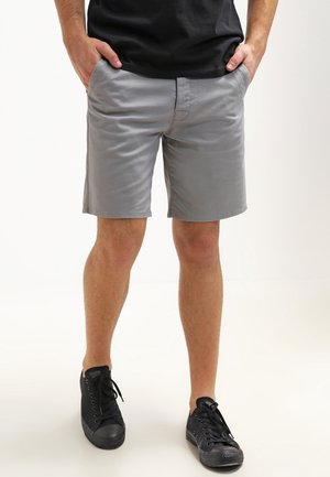 Mann steht mit den Händen in den Taschen, trägt graue Shorts, schwarzes T-Shirt und schwarze Turnschuhe auf weißem Hintergrund.