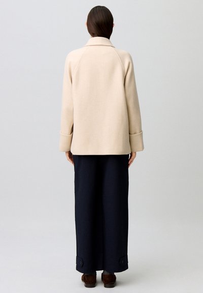 Donna in piedi di spalle che indossa un cappotto beige con ampi polsini, una lunga gonna nera con dettagli a bottoni e scarpe marroni su uno sfondo semplice.