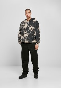 Un tânăr stă îmbrăcat cu un hanorac negru și bej cu model tie-dye, pantaloni negri largi și teniși negri, pe un fundal luminos simplu.