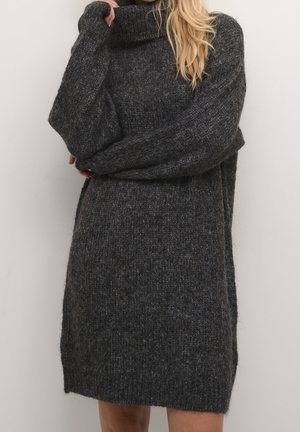 Woman wearing a dark gray chunky knit turtleneck sweater dress with long sleeves, standing against a plain light wall.