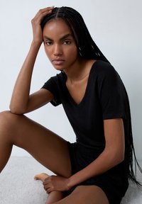 T-shirt nera a V, realizzata in tessuto leggero, con maniche corte e vestibilità comoda, indossata con pantaloni corti scuri su uno sfondo neutro.
