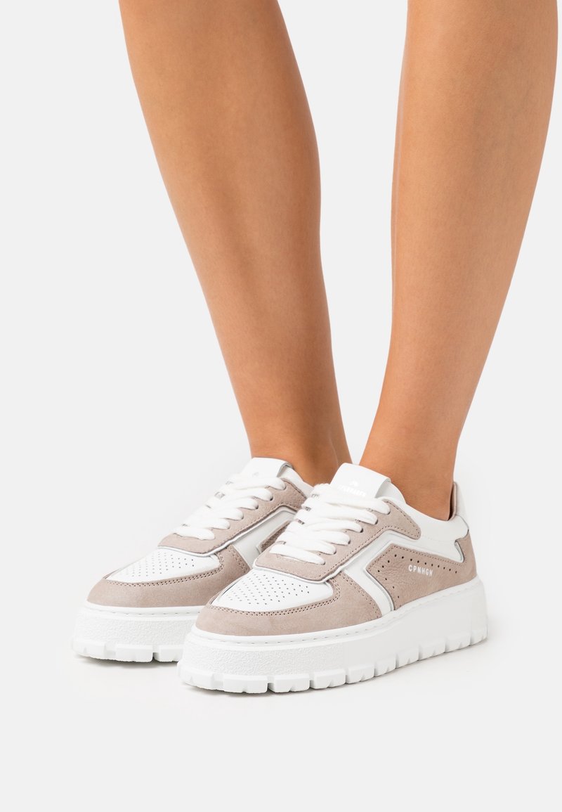 Jambes portant des baskets compensées en daim blanc et beige avec des détails perforés et des semelles blanches épaisses et texturées sur un fond clair.