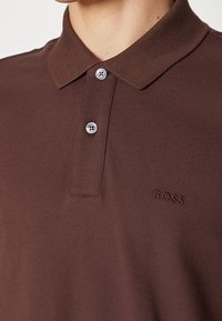 Gros plan d'un polo marron avec deux boutons argentés et le logo BOSS brodé sur la poitrine.