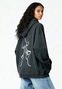 Donna con lunghi capelli ricci, che indossa un hoodie nero oversize con un'illustrazione astratta bianca di un volto sulla schiena e jeans blu.